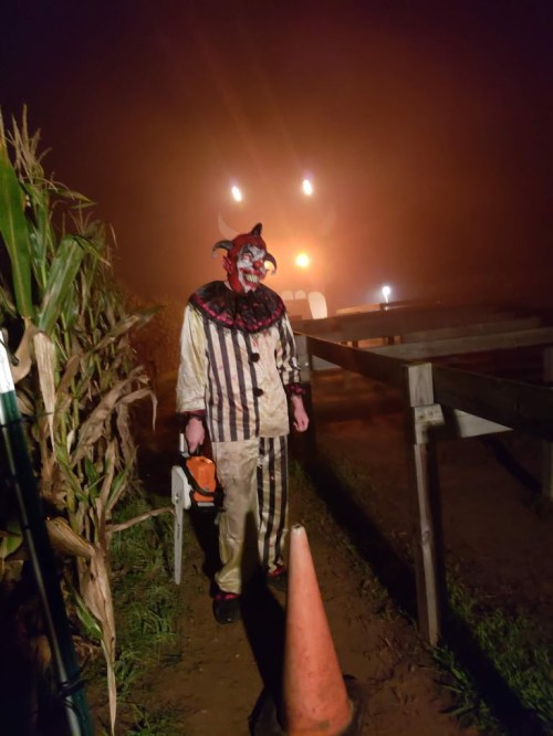 A creepy clown in a striped outfit stands in a foggy cornfield, holding a chainsaw near a traffic cone.