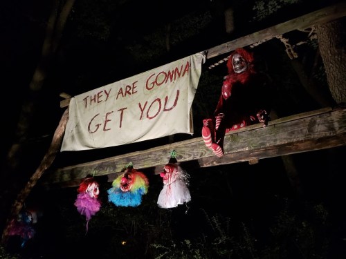A creepy scene in the woods featuring a sign that says "They are gonna get you" and hanging clown decorations.