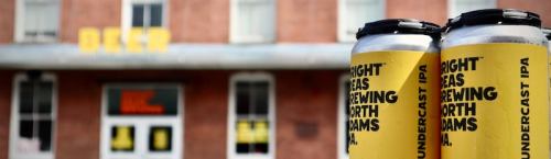 Two yellow cans of North Seas Brewing's Undercast IPA in front of a brick building with "DEEP" signage.