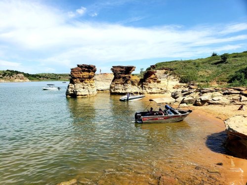 A serene lakeside scene with rocky formations, boats anchored near the shore, and a clear blue sky.