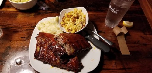 A plate with BBQ ribs, creamy mashed potatoes, and macaroni and cheese, alongside a glass of water and a lemon wedge.