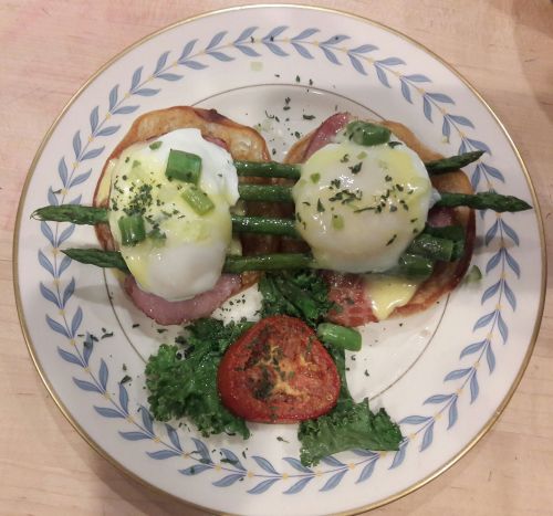 A plate of eggs Benedict with poached eggs, asparagus, ham, and a baked tomato, garnished with herbs and greens.