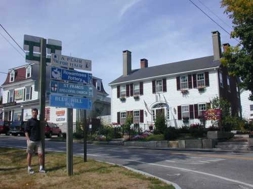 A man stands near a street sign with directions, in front of a white building with flower boxes and a welcoming entrance.