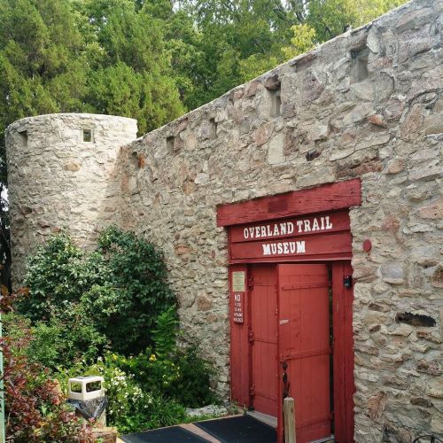 Stone building with a red door labeled "Overland Trail Museum," surrounded by greenery and a circular tower.