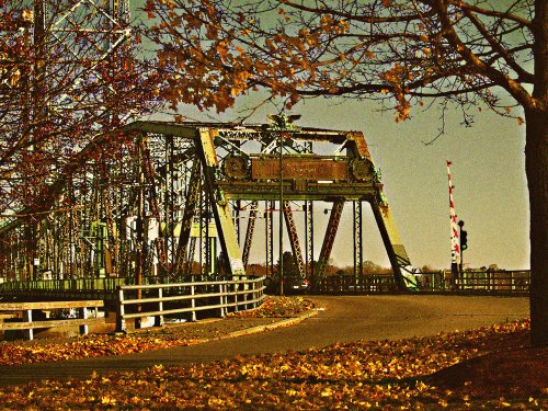 A vintage bridge with a metal structure, surrounded by autumn leaves and trees, under a clear sky.