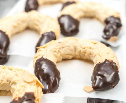 A close-up of crescent-shaped cookies dipped in chocolate and sprinkled with almond slices on parchment paper.