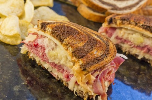 A close-up of a sliced sandwich with layers of meat and sauerkraut, served with potato chips in the background.