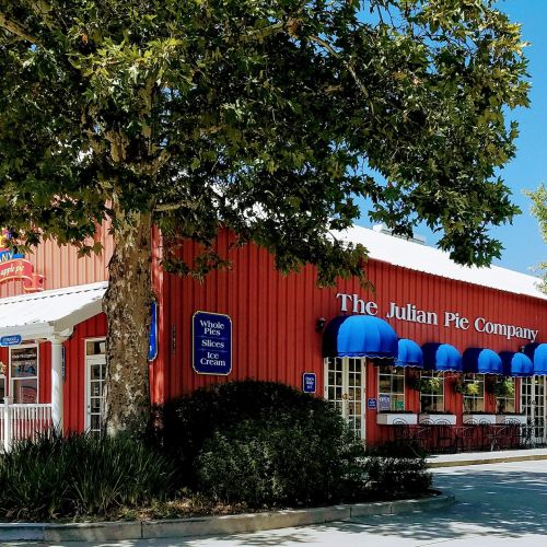 A red building with blue awnings, featuring "The Julian Pie Company" sign and outdoor seating under a tree.