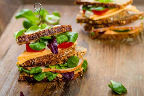 A stacked sandwich with layers of cheese, tomato, and greens on whole grain bread, served on a wooden board.