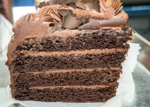 A slice of rich chocolate cake with layers of dark chocolate frosting and decorative chocolate shavings on top.