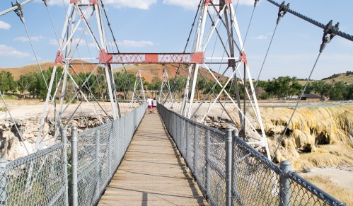 A wooden suspension bridge with metal supports, leading to a grassy area, under a blue sky with scattered clouds.