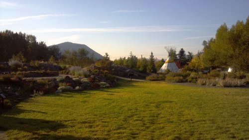 A serene landscape featuring a grassy area, trees, and a teepee, with mountains in the background under a clear sky.