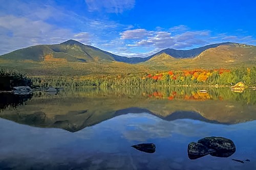 Scenic view of mountains reflecting in a calm lake, surrounded by colorful autumn foliage under a blue sky.