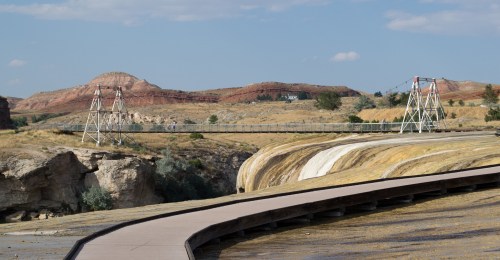 A suspension bridge spans a rocky landscape with hills in the background under a clear blue sky.