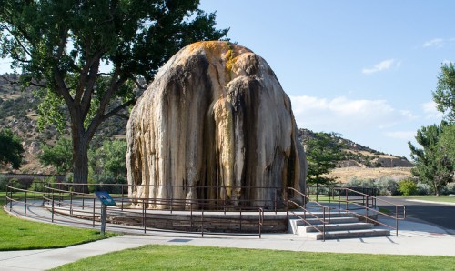 A large, mineral-rich rock formation surrounded by grass and trees, with a viewing platform and blue sky in the background.