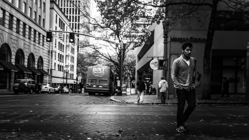 A man walks alone on a city street, surrounded by buildings and parked vehicles, in a black and white scene.