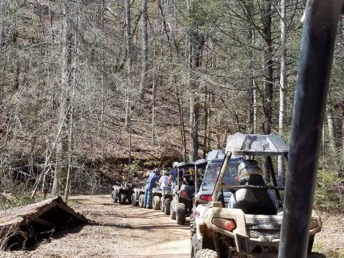 A line of off-road vehicles on a dirt trail surrounded by trees in a wooded area.