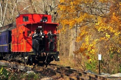 A red caboose on a train track surrounded by autumn trees, with two people standing on the back.