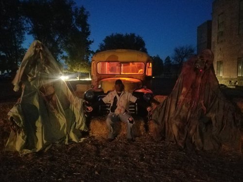 A spooky scene with two ghostly figures flanking a vintage yellow school bus at night, surrounded by hay bales.