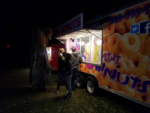 A food truck selling mini donuts at night, with two people ordering and a figure in a costume nearby.