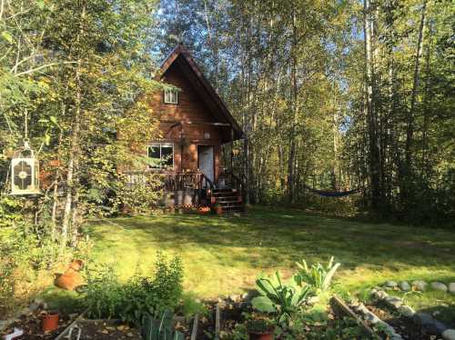 A cozy wooden cabin surrounded by trees, with a garden in front and a hammock hanging nearby.