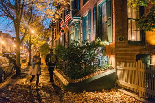A couple walks along a tree-lined street at dusk, with autumn leaves and historic brick buildings in the background.