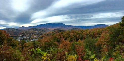 A panoramic view of mountains surrounded by vibrant autumn foliage under a cloudy sky.