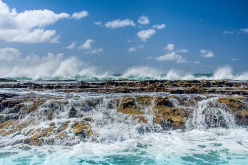 Waves crash against rocky shore under a bright blue sky with scattered clouds.