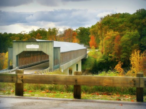 A covered bridge spans a river, surrounded by vibrant autumn foliage and a cloudy sky.