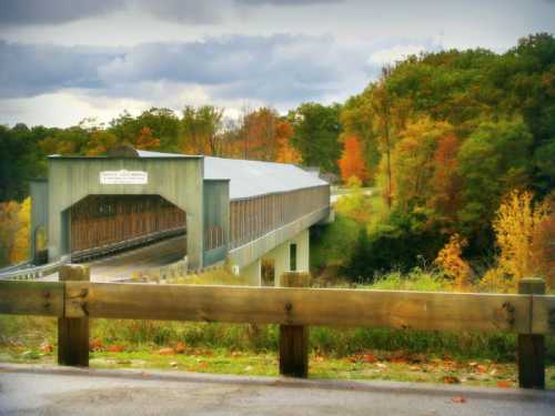 A covered bridge surrounded by colorful autumn trees under a cloudy sky. A wooden railing is in the foreground.