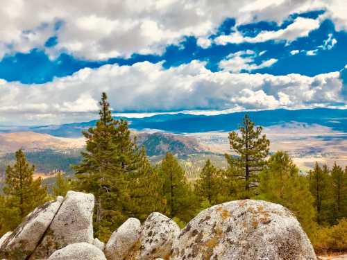 A scenic view of mountains and valleys, with lush green trees and large rocks under a cloudy blue sky.