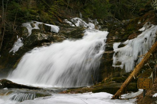 A frozen waterfall cascades over rocks, surrounded by ice and greenery, with a sign reading "This area closed to public."