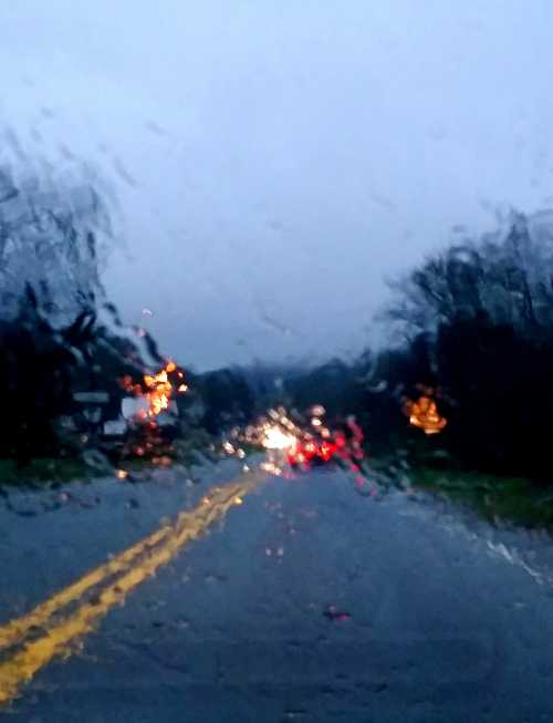 A rainy road scene with blurred lights and trees, viewed through a wet windshield.