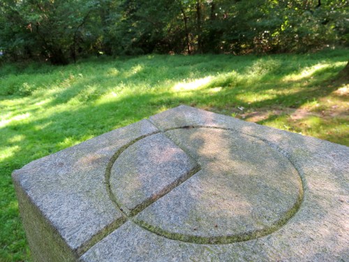 A stone pedestal with a circular carving on top, surrounded by lush green grass and trees in the background.