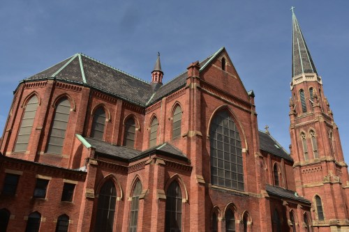 Historic red brick church with tall spire and large stained glass windows under a clear blue sky.