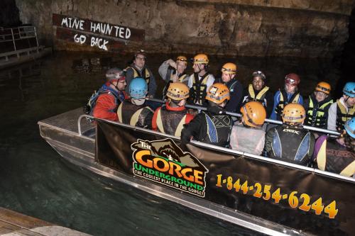 A group of people in helmets on a boat inside a cave, with a sign reading "MINE HAUNTED GO BACK" in the background.