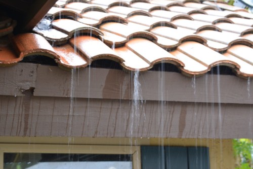 Water drips from a tiled roof, cascading down the edge onto a wooden surface below.