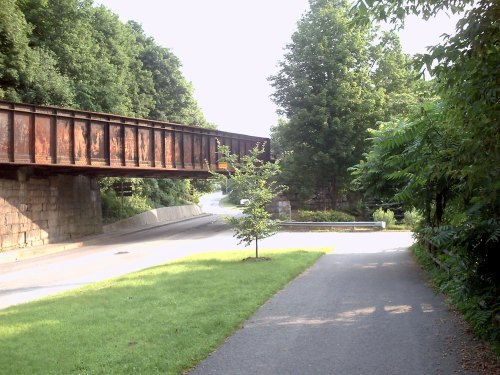 A pathway lined with greenery, leading to a bridge overhead, with a small tree and a road curving in the background.