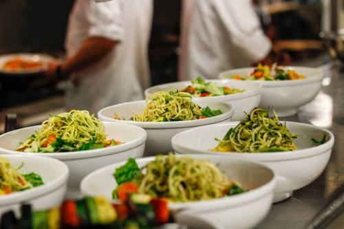 A row of white bowls filled with pasta and fresh vegetables, prepared in a busy kitchen. Chefs are visible in the background.