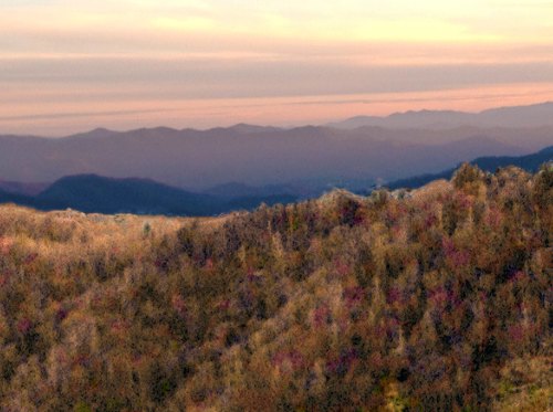A scenic view of rolling mountains at sunset, with soft hues of orange and purple in the sky and colorful foliage below.