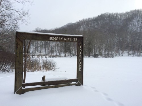 Snow-covered landscape at Hungry Mother State Park, with a wooden sign and a distant view of trees and a frozen lake.