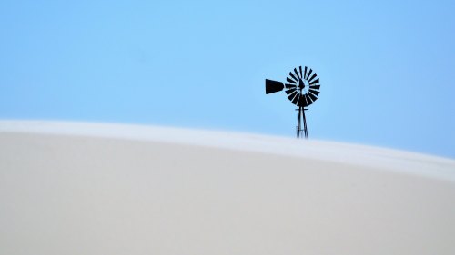 A silhouette of a windmill against a clear blue sky, partially obscured by a sandy dune.