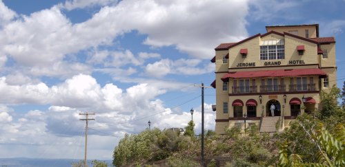 Historic Jerome Grand Hotel stands on a hillside under a cloudy sky, with power lines and greenery in the foreground.