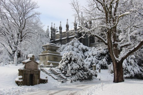 A snowy cemetery scene featuring ornate tombstones, trees covered in snow, and a grand building in the background.