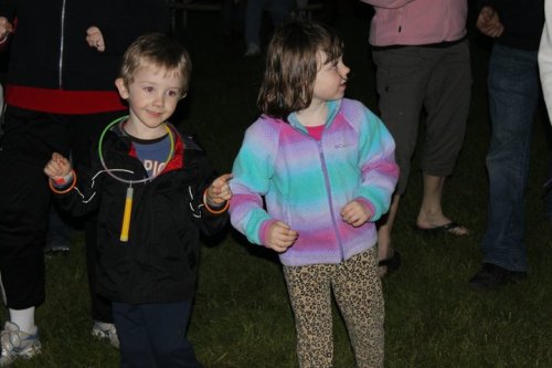 Two children dancing outdoors at night, one wearing a colorful jacket and the other in a black jacket with a glow stick.