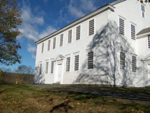 A white, two-story building with many windows, set against a blue sky and grassy area with scattered leaves.