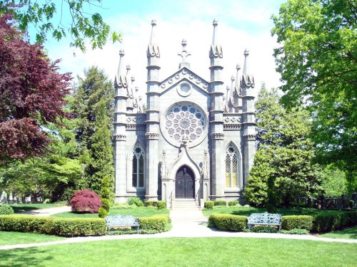 A gothic-style mausoleum surrounded by lush greenery and benches in a serene cemetery setting.