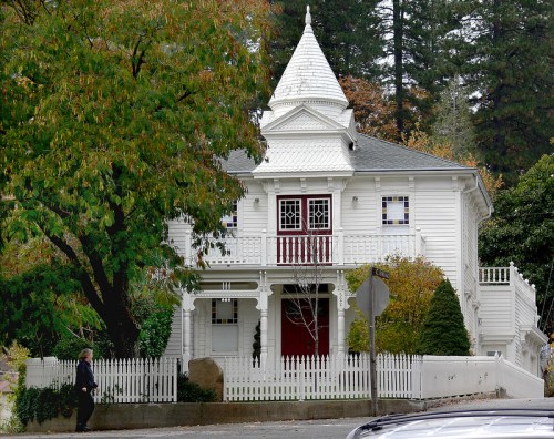 A charming white Victorian house with a turret, surrounded by trees and a white picket fence. A person walks by.