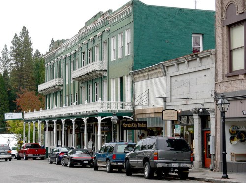 Historic buildings line a street in Nevada City, featuring a green hotel and various shops, with parked cars in view.
