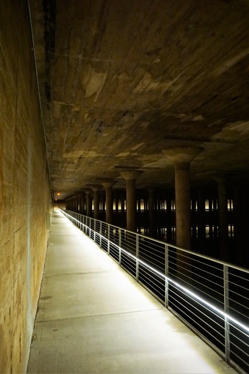 A dimly lit walkway under a concrete structure, with columns and a railing beside a calm body of water.